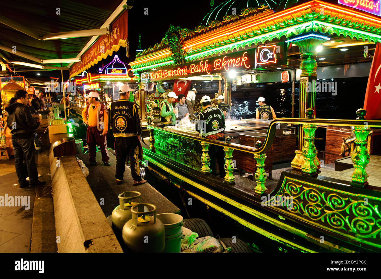 Restaurants flottants Balik Ekmek Eminonu Istanbul // ISTANBUL, Turquie — les bateaux éclairés balik ekmek servent des sandwichs de poisson traditionnels aux clients le long du front de mer d'Eminonu, près du pont de Galata. Ces restaurants flottants, connus localement sous le nom de balıkçı tekneleri, proposent du maquereau grillé servi dans du pain blanc avec des oignons et de la laitue comme nourriture de rue populaire. Les bateaux richement décorés présentent des motifs de style ottoman et restent amarrés en permanence le long du front de mer historique de la Corne d'Or. Eminonu sert de terminal de ferry majeur et de quartier commercial reliant les côtés européens et asiatiques de ITS Banque D'Images