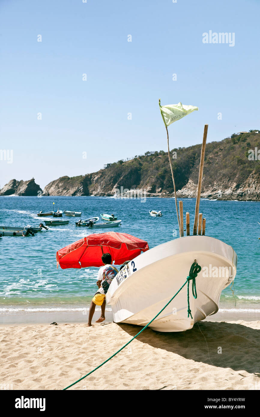 Bateau de pêche échoué sur la plage principale de la Mexican boy sitting on stern sous parapluie rouge à la baie d'azur à Puerto Angel Banque D'Images