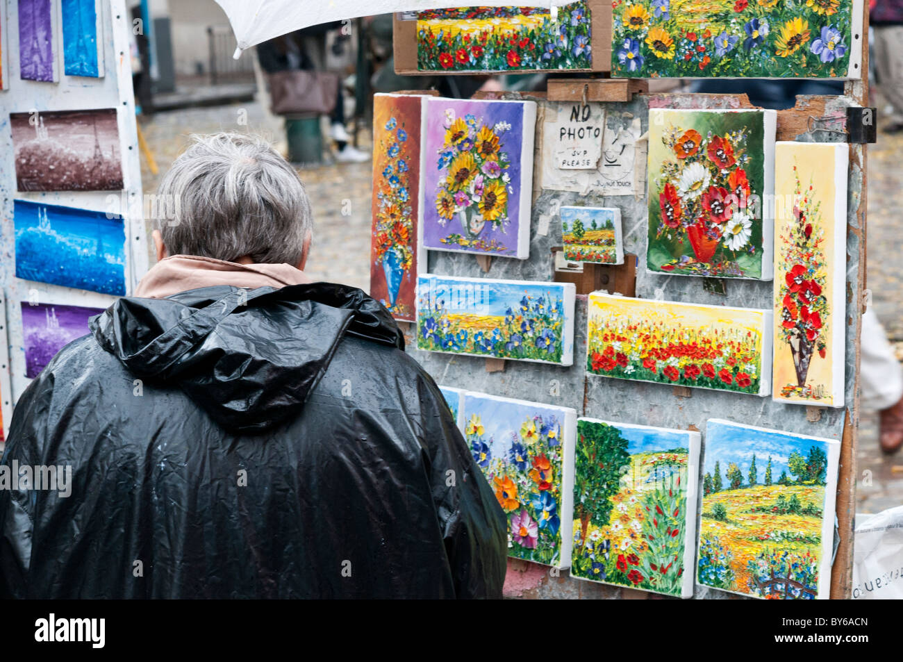 Exposition de peintures de rue de Montmartre Paris France // PARIS, France — Un visiteur observe une exposition extérieure de peintures, représentant des arrangements floraux et des représentations de la Tour Eiffel, d'un artiste de rue de Montmartre. Ce quartier emblématique parisien, en particulier la place du Tertre, est réputé pour sa communauté dynamique d’artistes de rue et son patrimoine bohème. Le charme historique et l'héritage artistique de Montmartre en font une destination populaire pour les touristes et les amateurs d'art. La région est connue pour ses chemins pavés et ses bâtiments historiques, contribuant à son caractère unique. Situé sur a la Banque D'Images