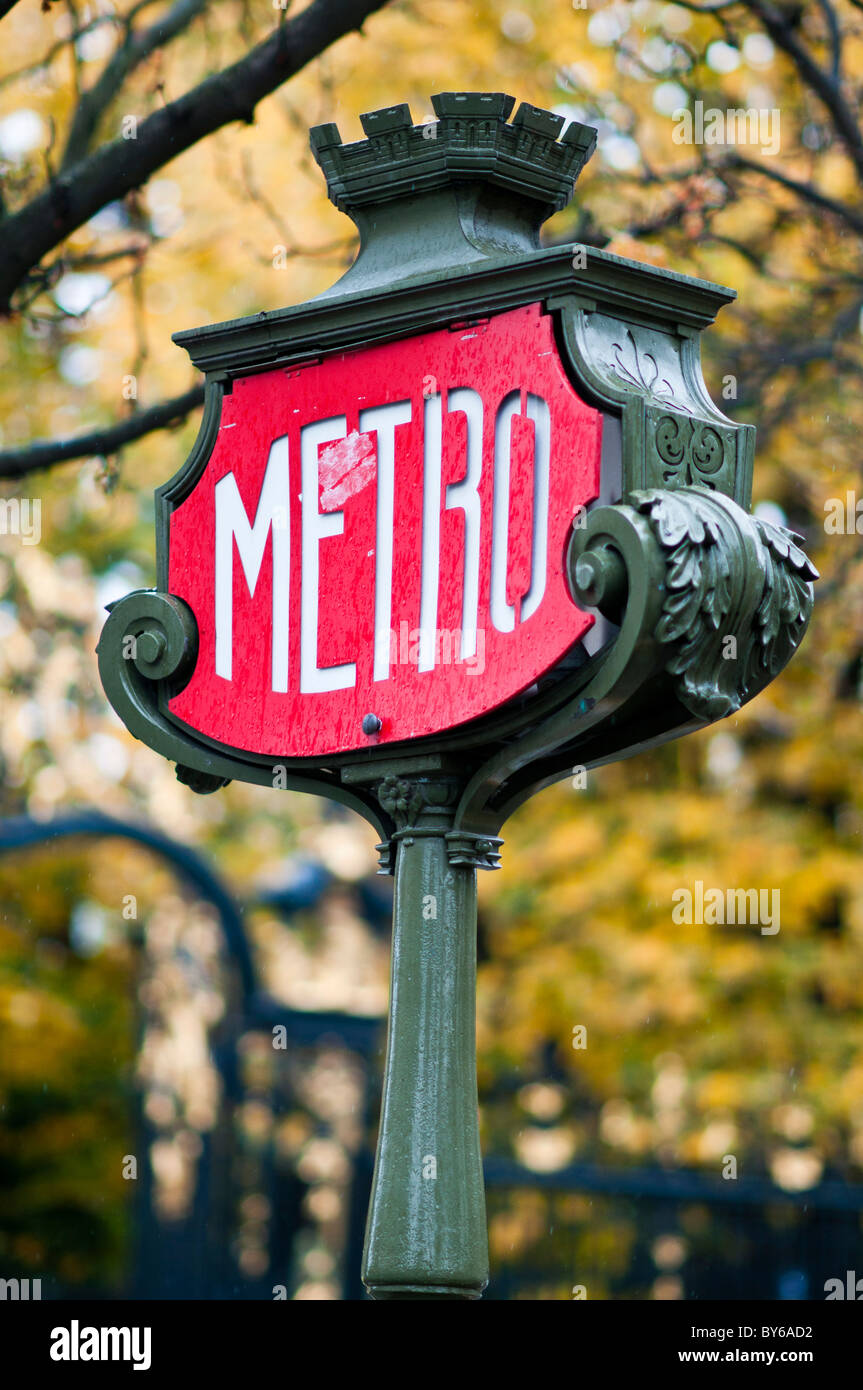 Panneau de métro Paris orné de fonte verte avec toit crénelé Paris France // PARIS, France — un panneau de métro Paris en fonte verte orné, avec un panneau de MÉTRO rouge et blanc distinctif et un toit crénelé, marque l'entrée d'une station de MÉTRO. Ces enseignes classiques sont un symbole emblématique du système de transport en commun parisien, exploité par la Régie autonome des Transports Parisiens (RATP). Le design de l'enseigne comprend des volutes décoratives et un sommet crénelé, caractéristiques de l'infrastructure urbaine historique de la ville. L'arrière-plan montre un feuillage automnal flou, suggérant un rai Banque D'Images