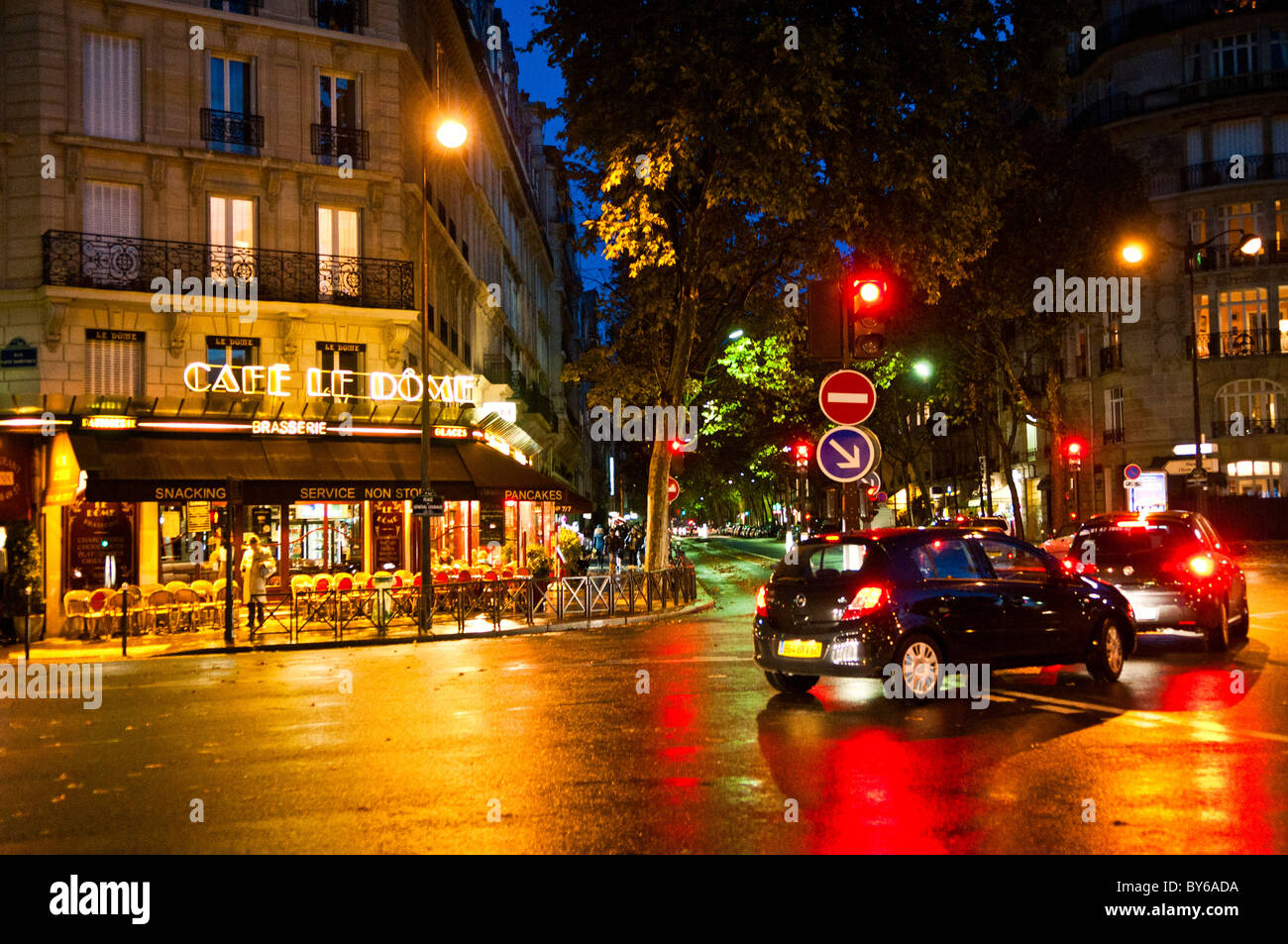Café Le Dôme Pavé scintillant Soirée parisienne 14e Arrondissement Paris France | PARIS, France — Le Café Le Dôme brille au-dessus d’une intersection trempée de pluie sur le Boulevard du Montparnasse dans le 14e arrondissement, son enseigne néon de brasserie se reflétant sur le pavé mouillé en dessous. Ouvert à la fin du XIXe siècle, le café est devenu un point de rencontre pour les artistes et écrivains de Montparnasse du début du XXe siècle et reste une brasserie en activité aujourd’hui. Les feux de circulation et les voitures qui passent strient de rouge la rue scintillante au crépuscule. Banque D'Images