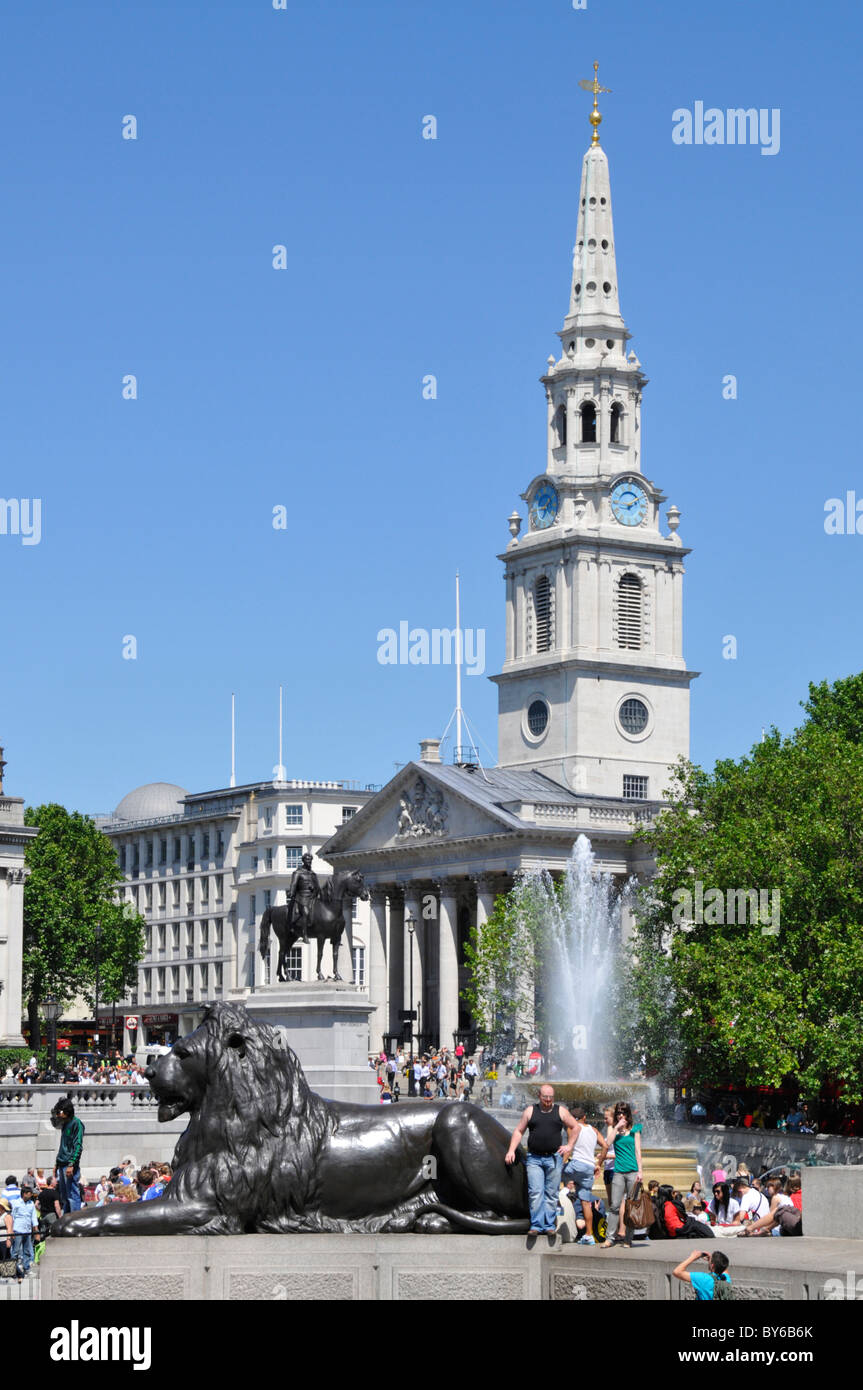 Les touristes à côté de l'eau de fontaine et le lion de bronze monumental à Trafalgar Square avec l'église anglicane St Martin-in-the-Fields et la flèche Londres Royaume-Uni Banque D'Images