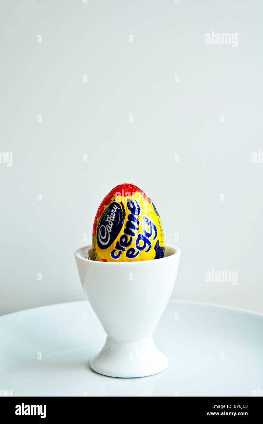 Un Cadbury Creme Egg in a white egg cup sur une plaque blanche avec fond uni. Banque D'Images