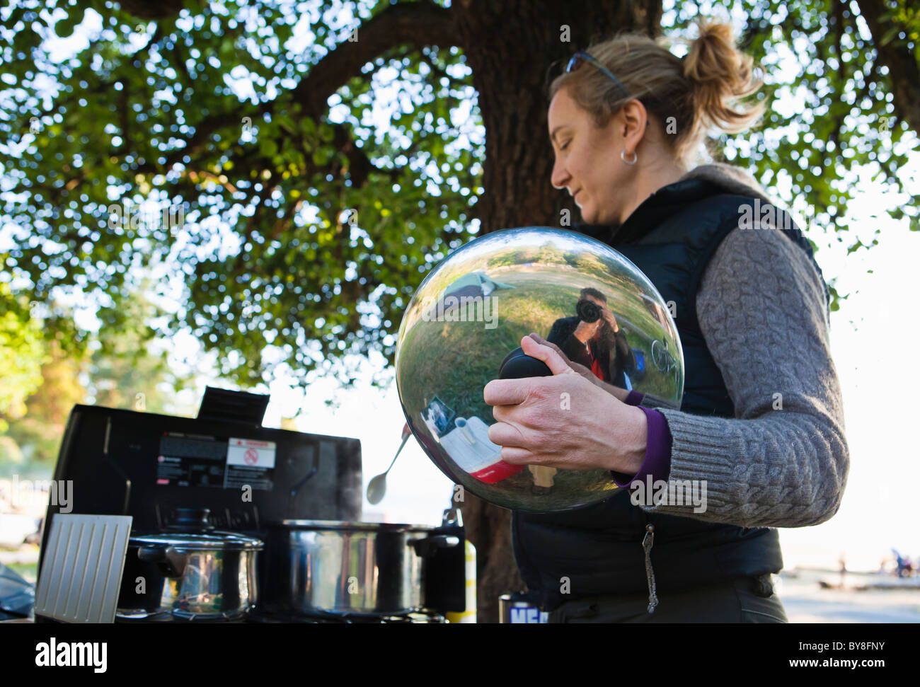 Un homme de prendre une photo reflète dans la casserole couvercle tenu par une femme la cuisson à l'extérieur sous un arbre. Banque D'Images