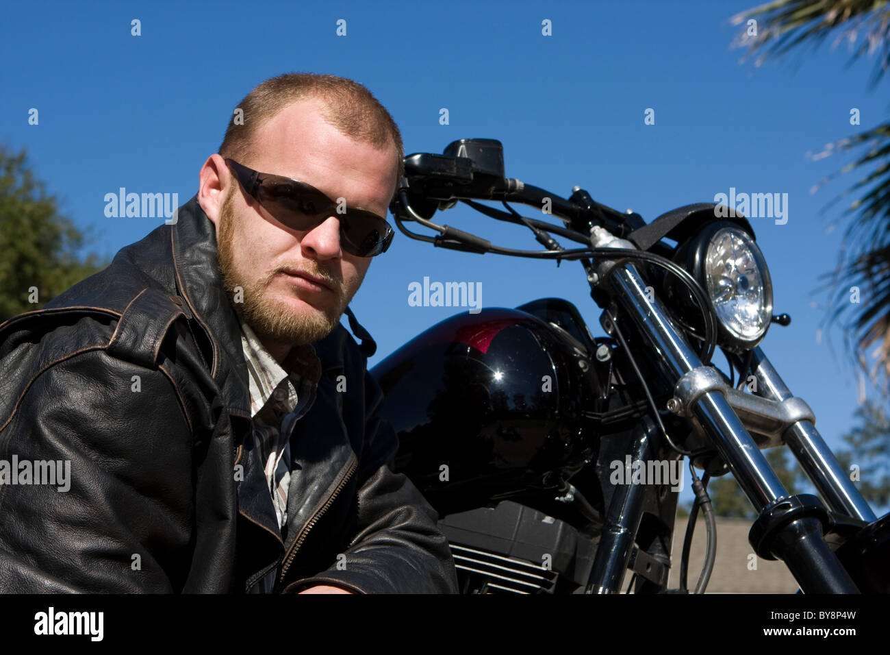 Biker pose devant sa moto en veste en cuir noire avec un regard sérieux. Banque D'Images