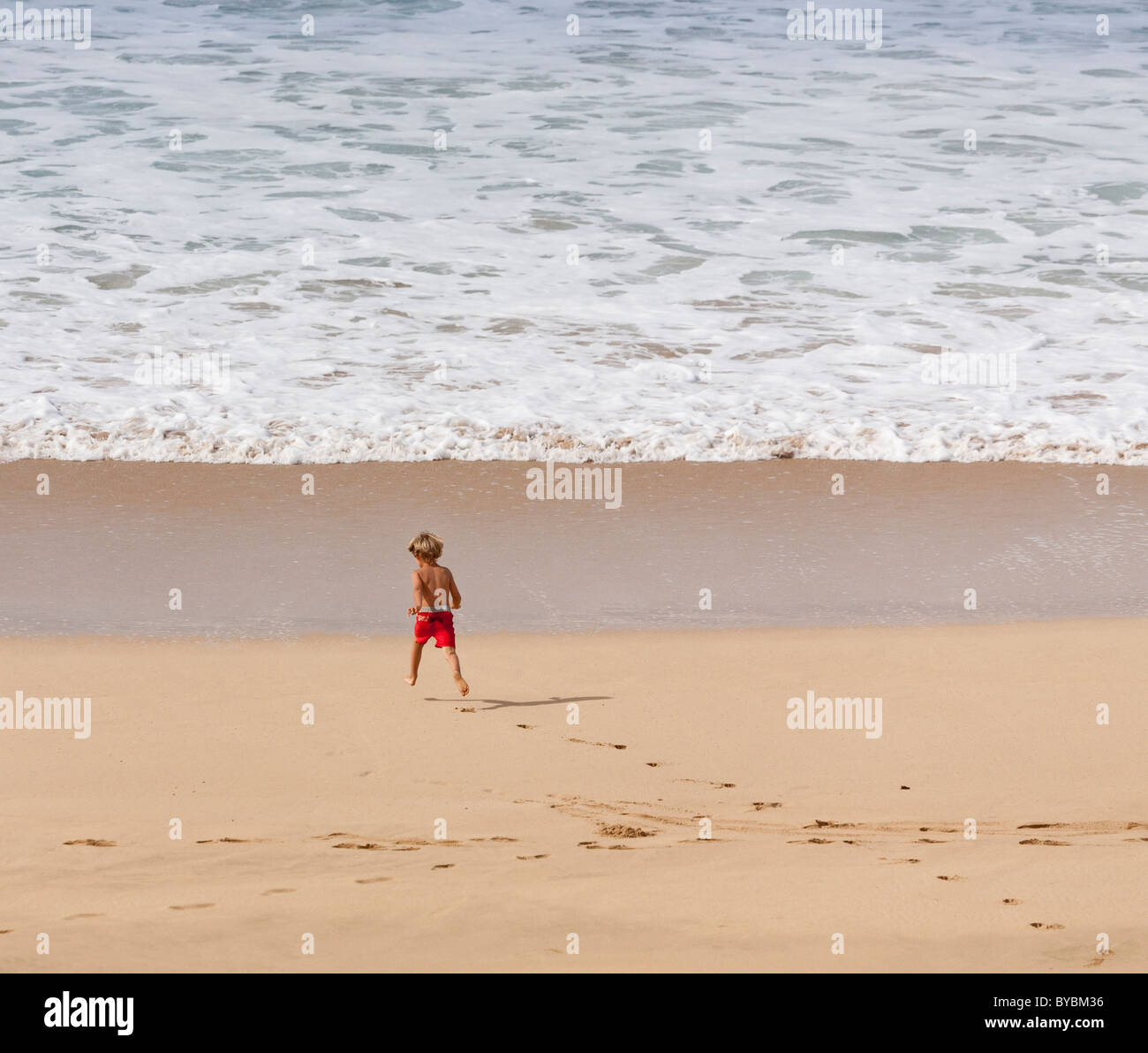 L'exécution du bord. Un jeune garçon en rouge les lignes de bain coule Polihale plage vers le bord de l'eau. Banque D'Images