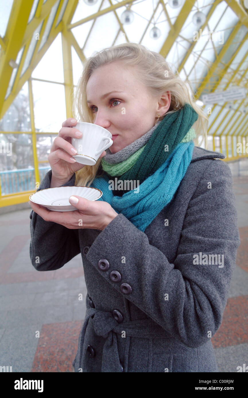 La jeune fille blanche avec des cheveux longs ondulés dans un capuchon gris et un manteau chaud boissons dans une tasse de thé dans le pied Pushkinsky pont, Banque D'Images