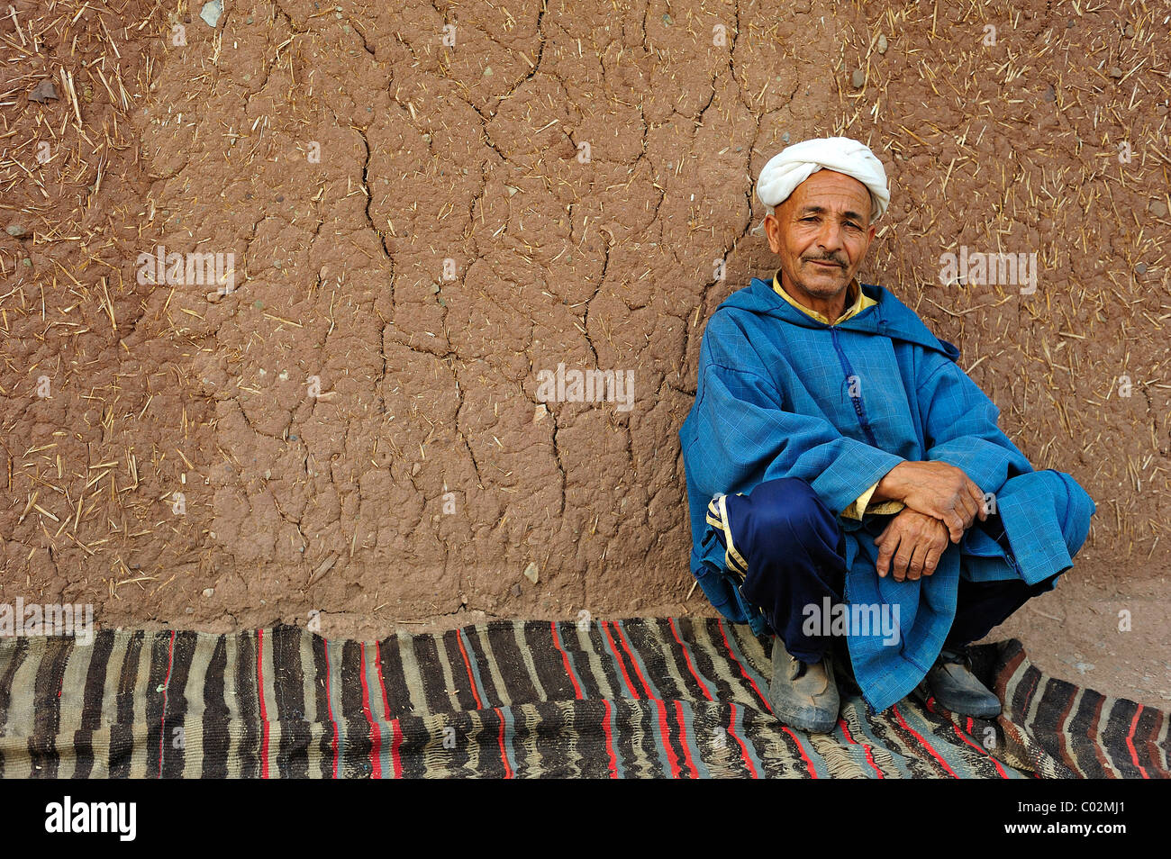 Portrait d'un homme âgé portant une Djellabah traditionnelle et un turban assis sur une couverture et appuyé contre le mur de boue Banque D'Images