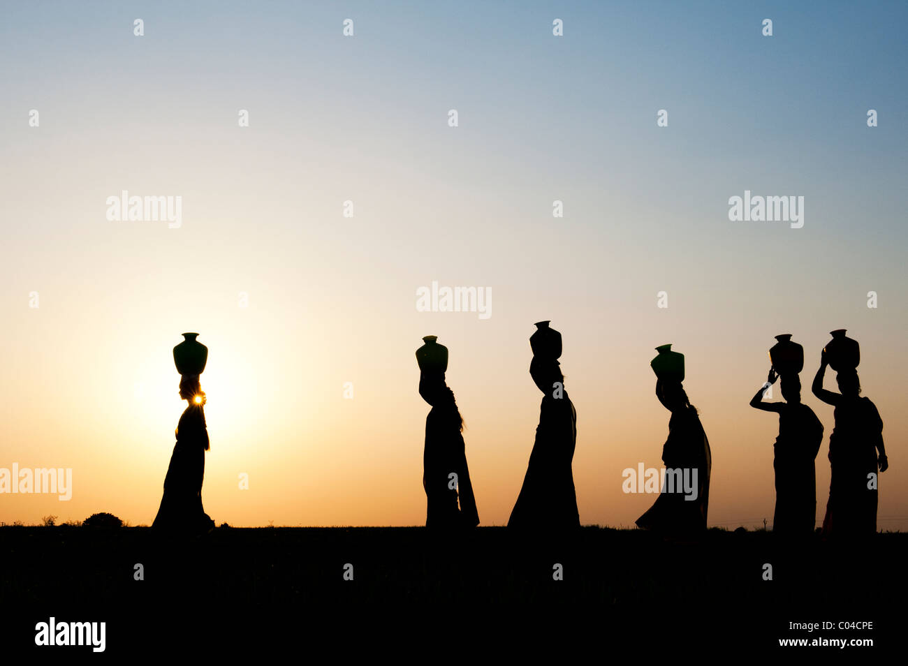 Groupe de femmes indiennes balade au coucher du soleil à transporter de l'eau sur leurs têtes des pots dans la campagne indienne. Silhouette Banque D'Images