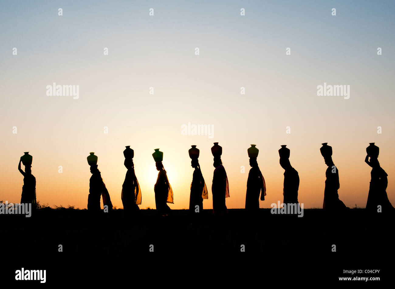 Groupe de femmes indiennes balade au coucher du soleil à transporter de l'eau sur leurs têtes des pots dans la campagne indienne. Silhouette Banque D'Images