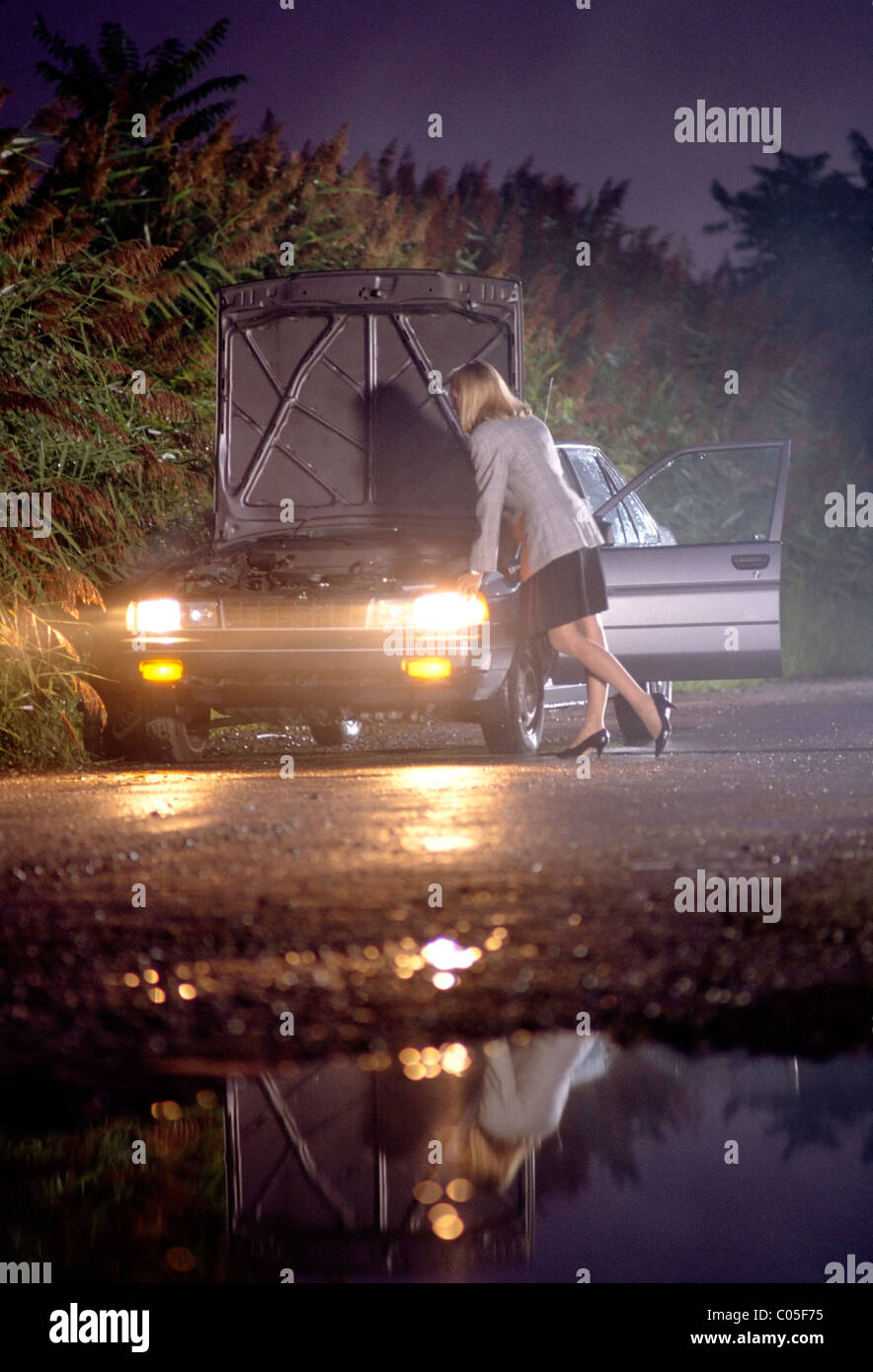 Femme vêtue de tenue de vérifications sous le capot de sa voiture en panne sur une nuit de brouillard de pluie. Banque D'Images