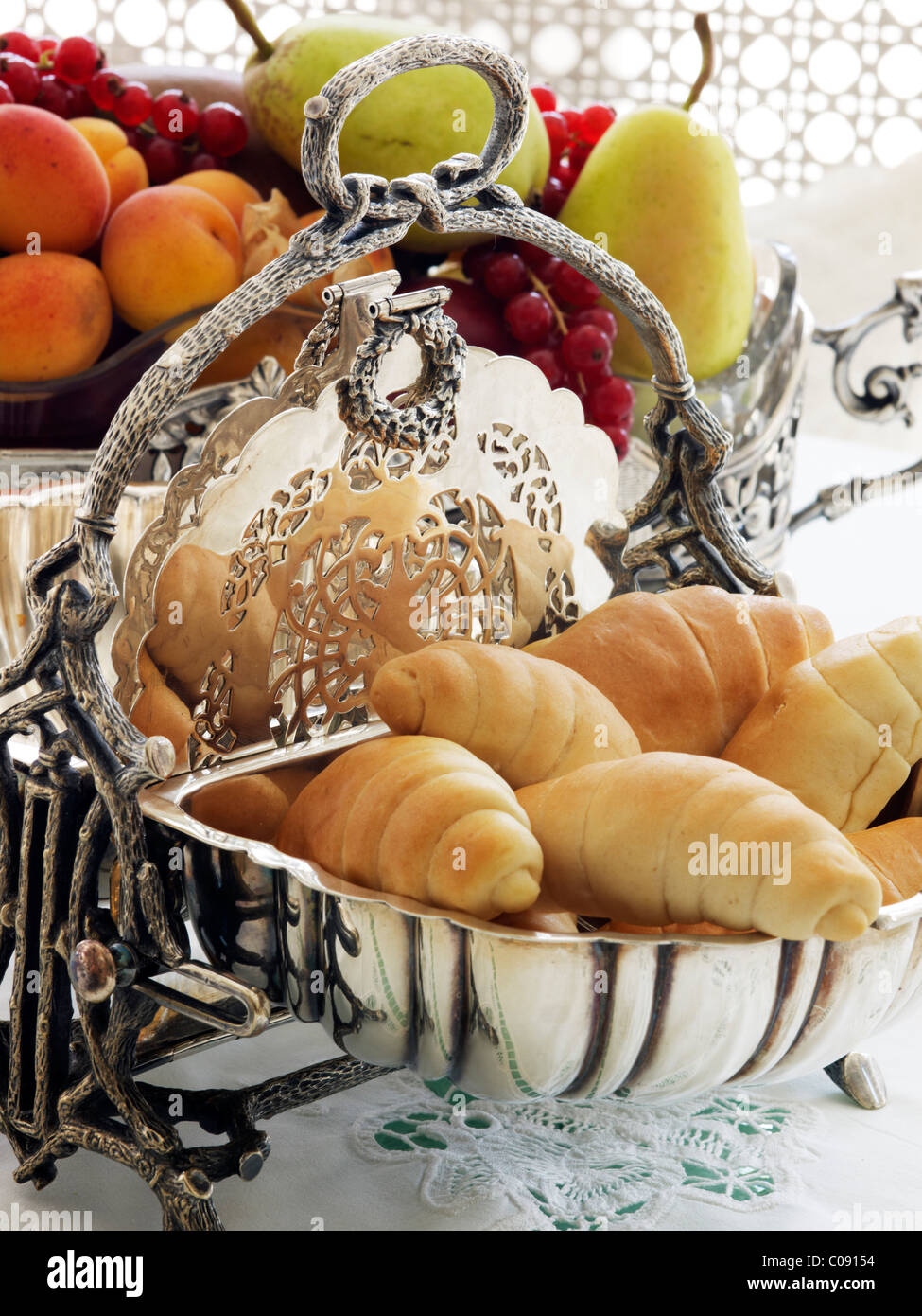 Argenterie ancienne panier à pain à charnière avec des croissants devant un bol de fruits sur une table de petit-déjeuner Banque D'Images