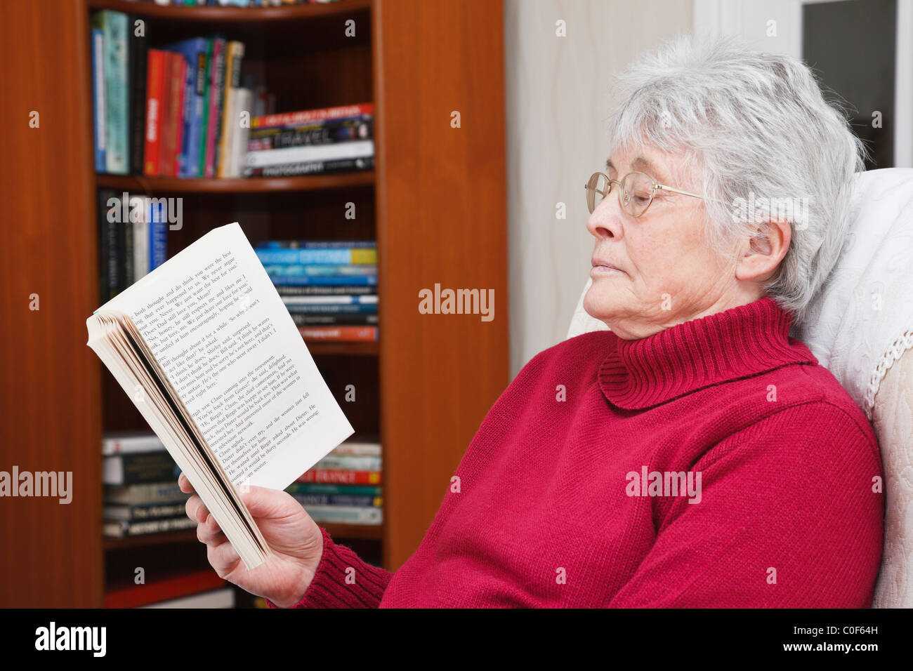 Quelqu'un femme âgée portant un cavalier rouge assis relaxant de lire un livre de poche dans un salon avec une bibliothèque derrière. UK Banque D'Images