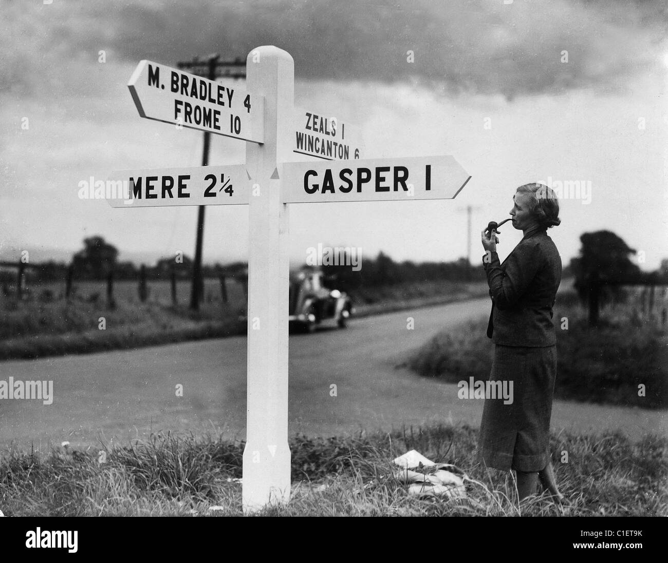 Dame fumant une pipe standing next to road sign post dans le Wiltshire en 1930. Banque D'Images
