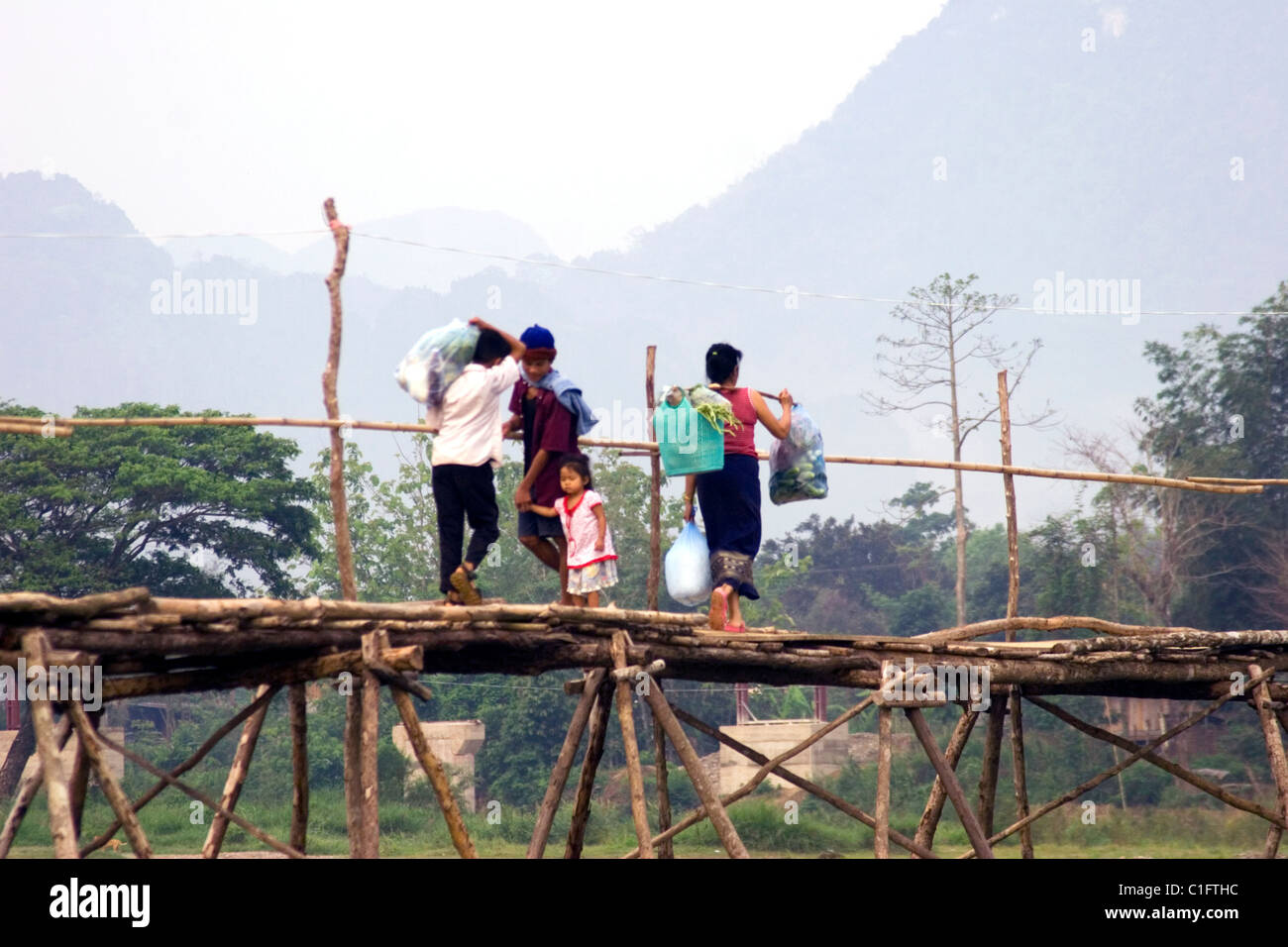 Un groupe de personnes traversent une rivière rural en marchant sur un pont en bois dans le Laos communiste. Banque D'Images