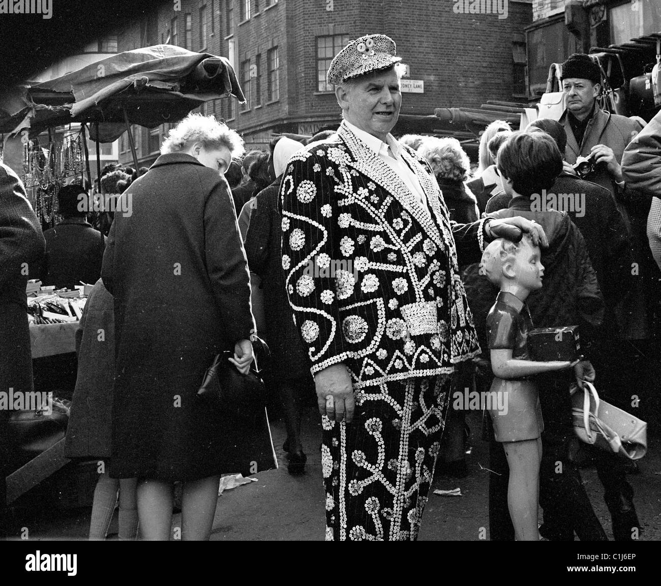 Années 1950, un roi nacré dans sa tenue décorée se tient dans un marché en plein air animé de l'East End avec sa main sur une boîte de charité Poor, Londres, Angleterre, Royaume-Uni. Banque D'Images