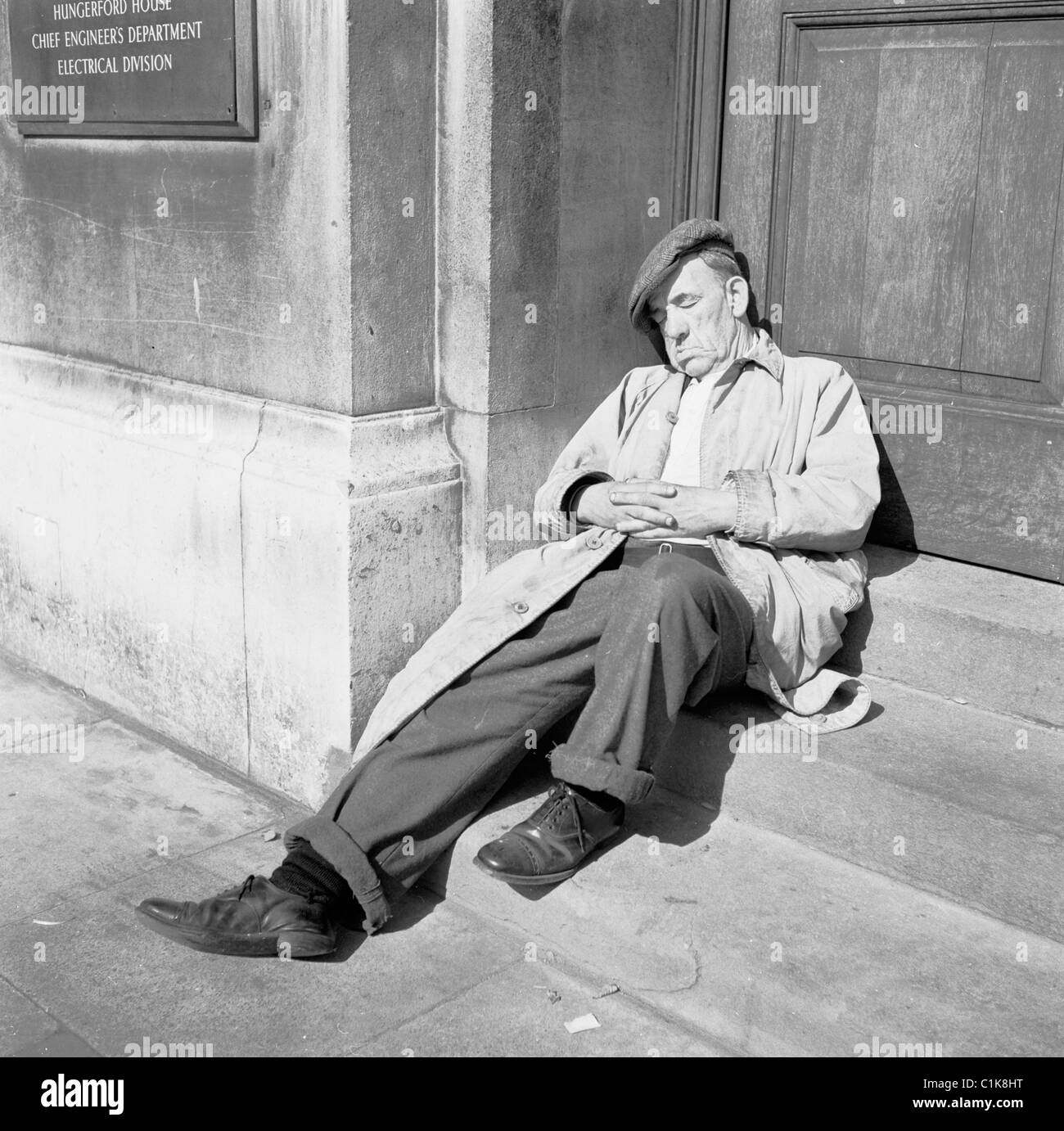 1950s, un vieil homme fatigué, dans un imperméable et une casquette plate, peut-être un clochard, ayant une sieste d'après-midi sur les marches de Hungerford House, Embankment, Londres. Banque D'Images