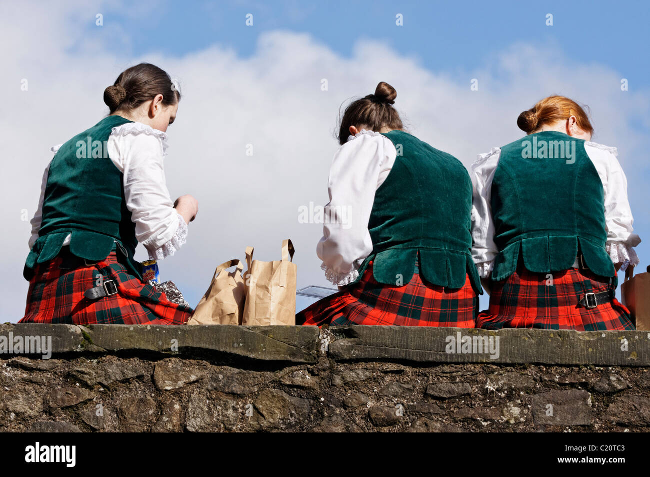 Trois jeunes filles portant des vêtements de danse des highlands assis en train de déjeuner sur un mur au château de Stirling, Scotland, UK. Banque D'Images