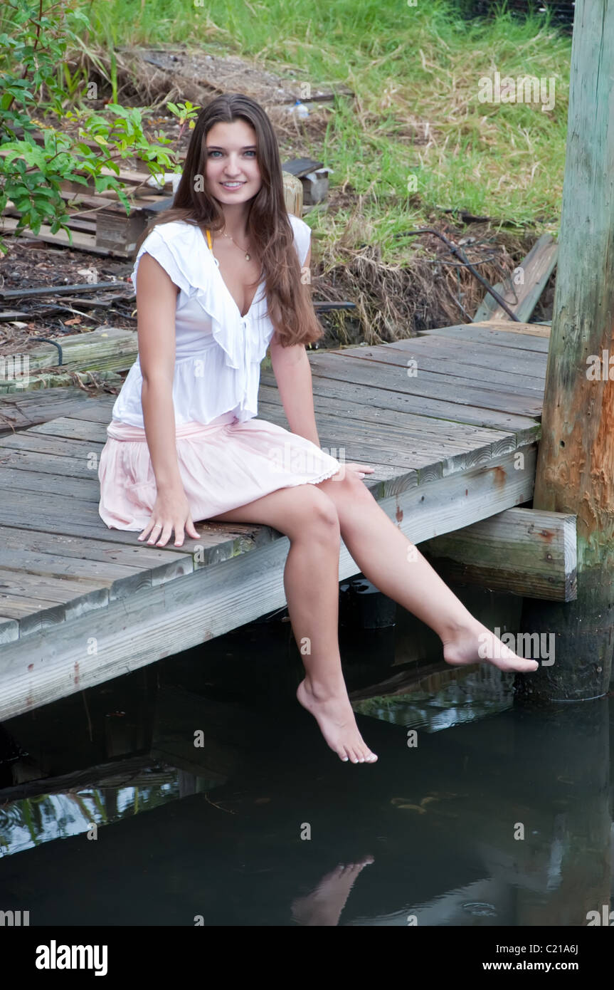 Jolie jeune fille assise sur un quai, les pieds ballants dans l'eau. Banque D'Images