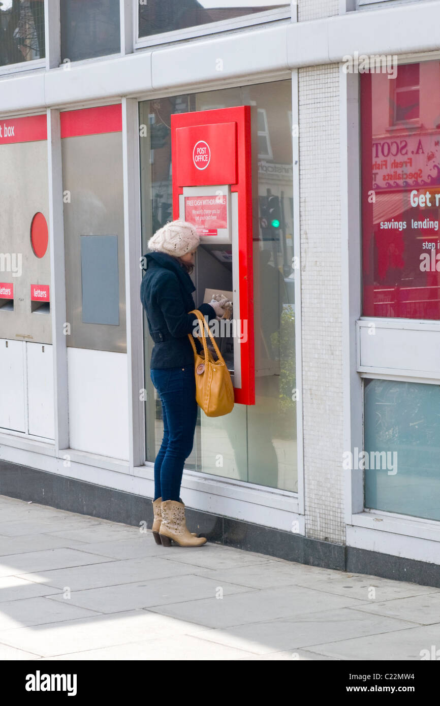 East Finchley , jolie jeune fille en bottes de cow-boy et blanc woolly hat tire de l'argent du bureau de poste distributeur automatique ou un trou dans le mur Banque D'Images