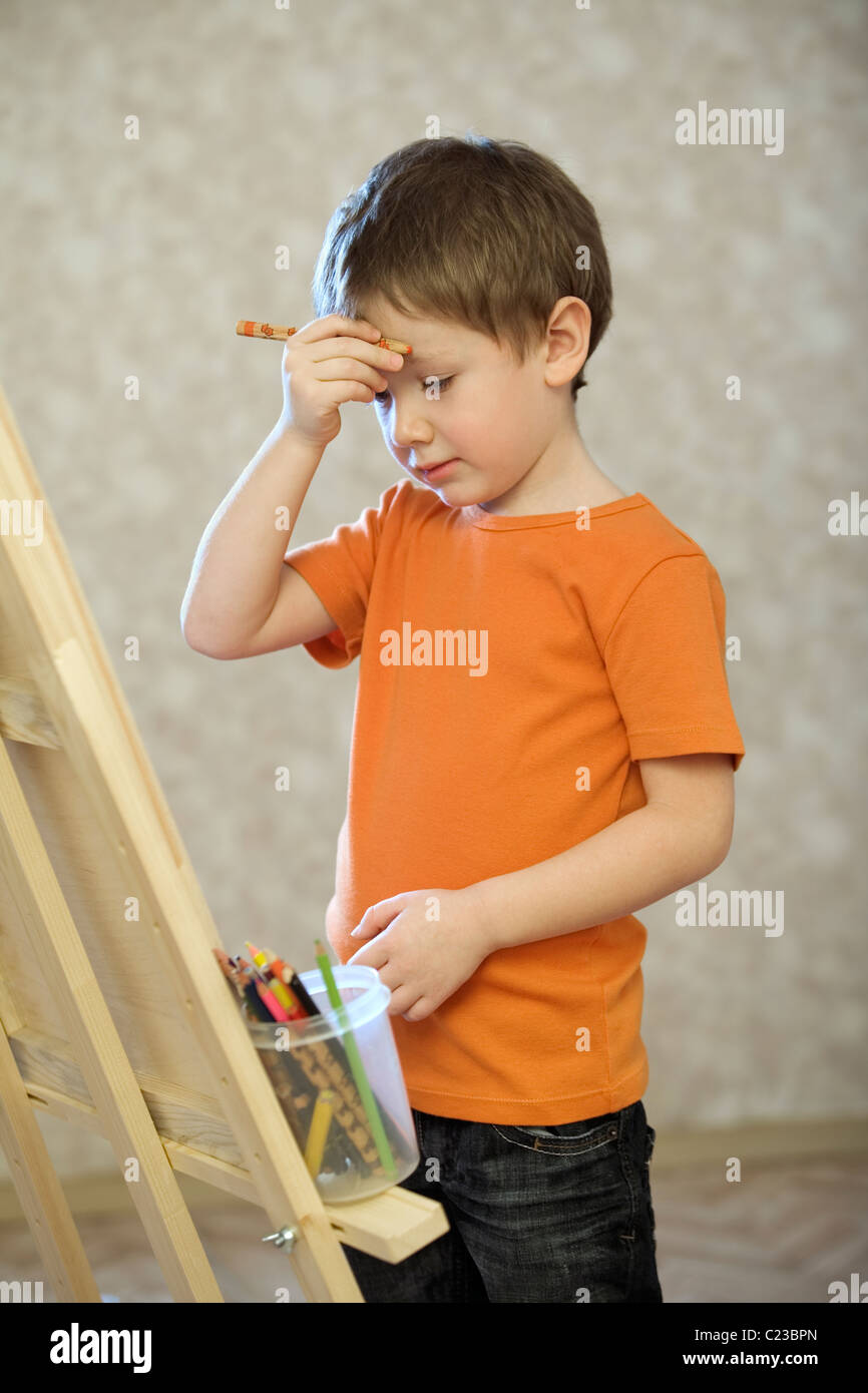 Un jeune garçon tenant la main avec un crayon en jusqu'à son visage:debout en face d'une toile avec un pot de crayons de couleur Banque D'Images