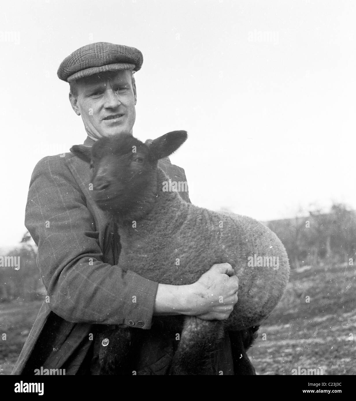 Années 1950, l'Angleterre. Un agriculteur détient un mouton noir dans ses bras dans ce tableau historique par J Allan l'argent comptant. Banque D'Images