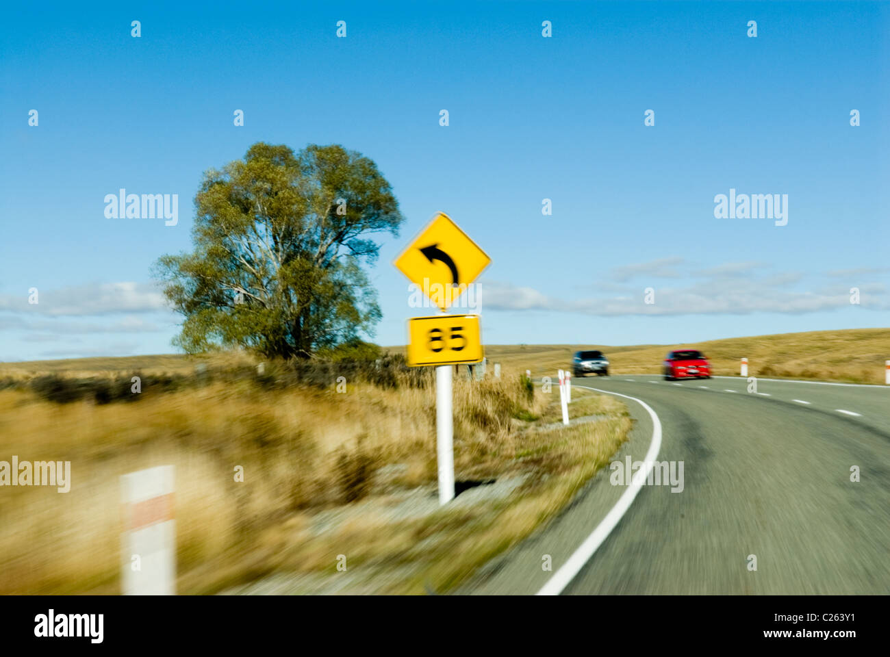 Effet de flou de mouvement - 85 km/h, virage gauche consultatif vitesse signe. L'approche des voitures rouge et argent. Banque D'Images