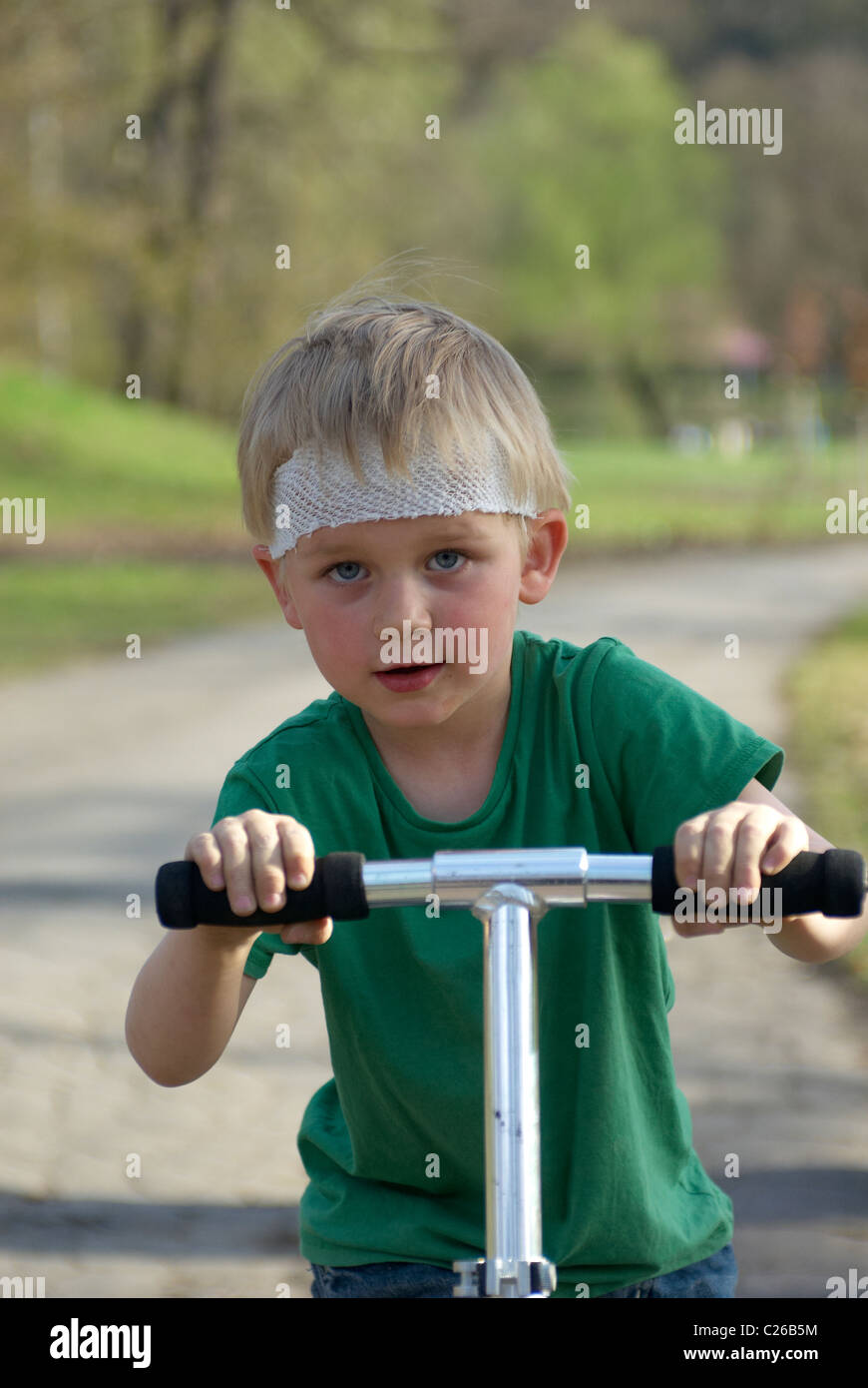 Un jeune enfant garçon blond sur un scooter dans parc avec tête blessée (bandage capeline) sans casque de sécurité Banque D'Images