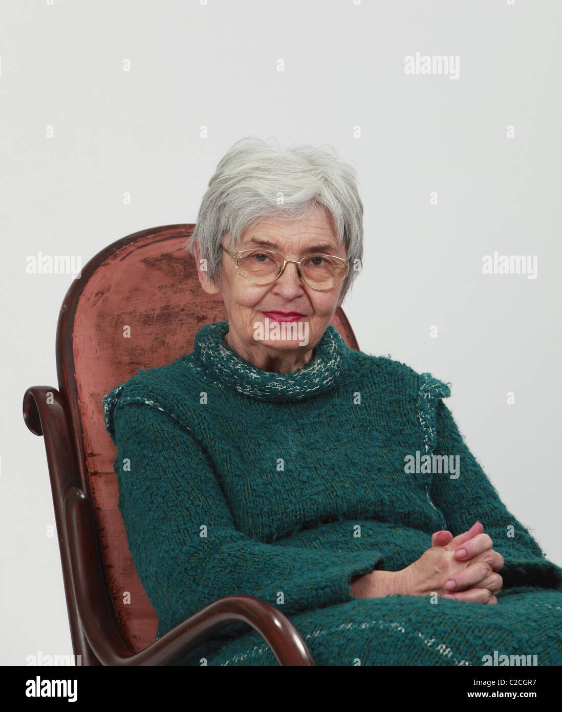 Portrait d'une vieille femme assise sur une chaise à bascule en bois avec les doigts, sur un arrière-plan gris. Banque D'Images