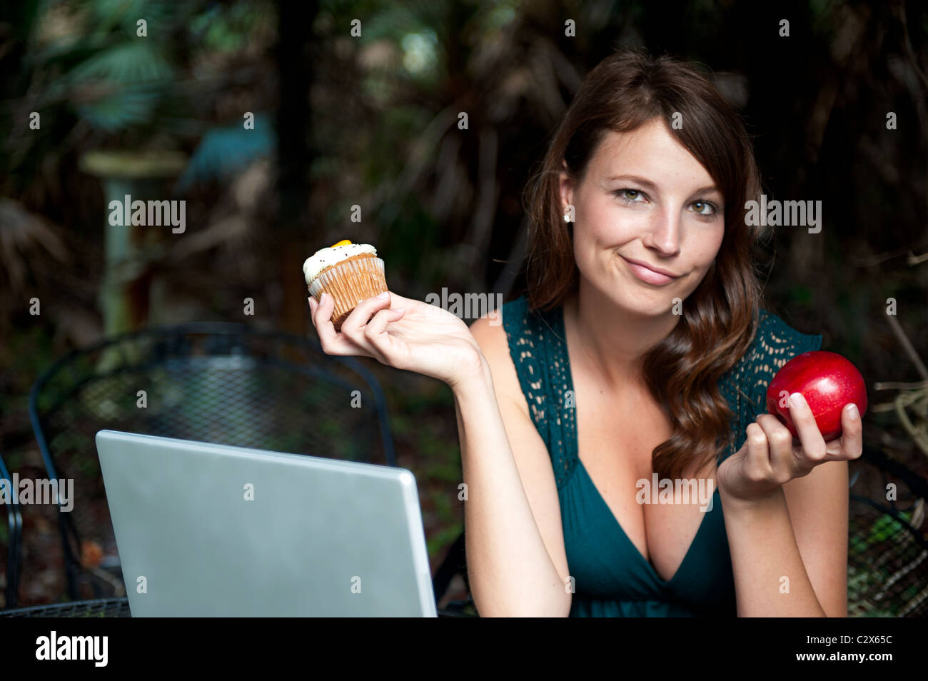 Jeune femme 30s devant ordinateur décide entre un petit gâteau et une pomme, la malbouffe contre l'alimentation saine. Banque D'Images