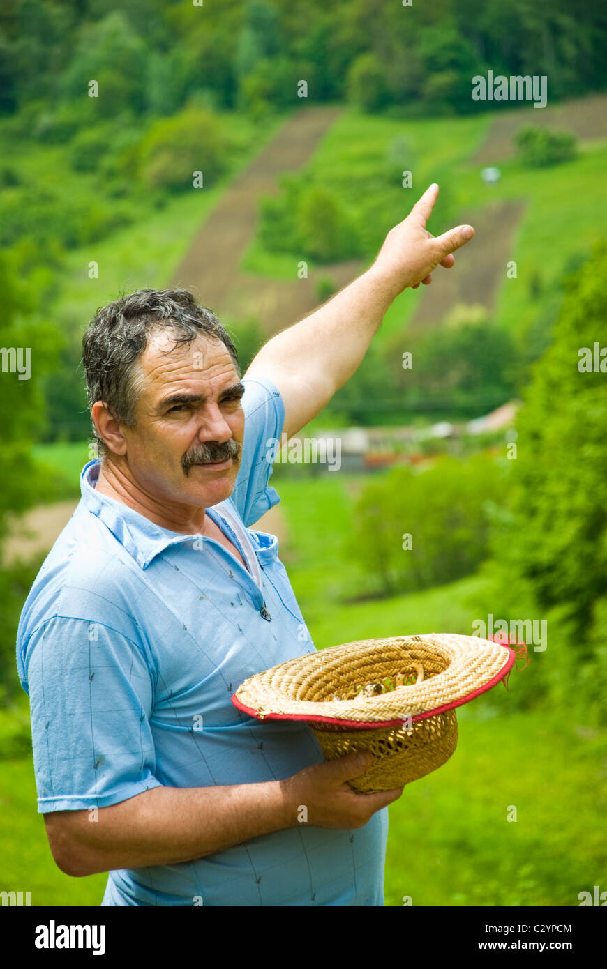 Farmer holding son chapeau de paille et montrant le labour des terres sur la campagne collines à un espace vert avec les terres cultivées et les forêts de nombreux Banque D'Images