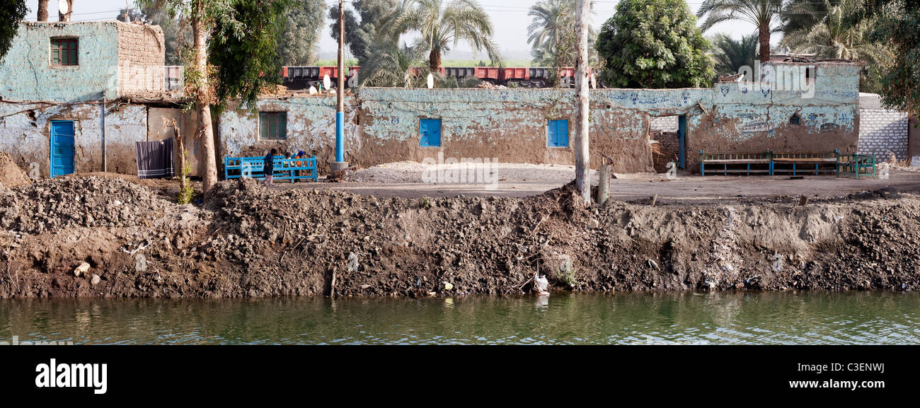 Maisons traditionnelles égyptiennes le long d'un canal boueux, banque avec bancs bleu et des enfants devant et derrière un train de marchandises, l'Égypte Banque D'Images