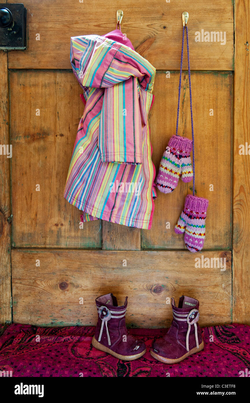 Les jeunes filles manteau et mitaines à rayures colorées suspendu par une cuisine en bois avec porte éraflée bottes sur le mat Banque D'Images