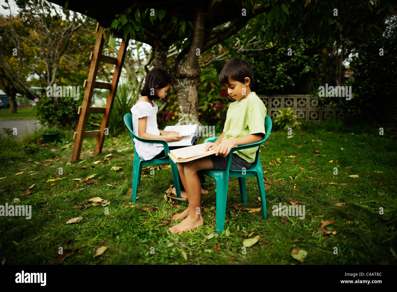 Garçon et fille s'asseoir sur des chaises en plastique des livres de lecture dans le jardin sous les arbres. Banque D'Images