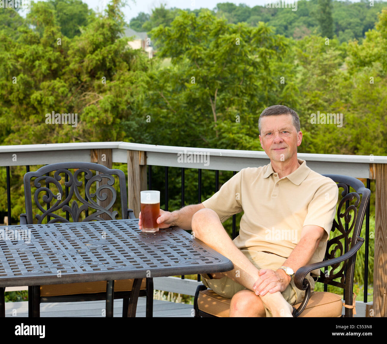 Homme d'âge moyen assis sur table de jardin en aluminium moulé sur le pont et de boire un verre de bière dans la cour arrière Banque D'Images