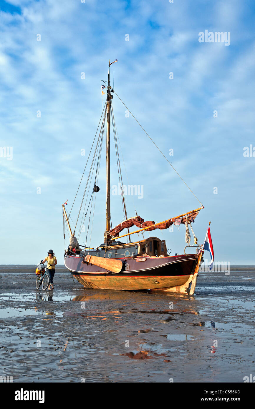 Les Pays-Bas, l'Île Ameland Nes, appartenant à des îles de la mer des Wadden. Bateau à voile sur la boue à plat dans le port. Femme et vélo. Banque D'Images