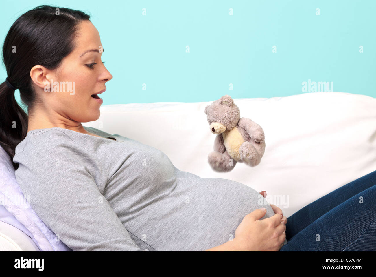 Photo d'une femme enceinte allongée sur un canapé aux prises avec son bébé premier kick, regard de surprise sur son visage comme le petit ours en peluche être Banque D'Images