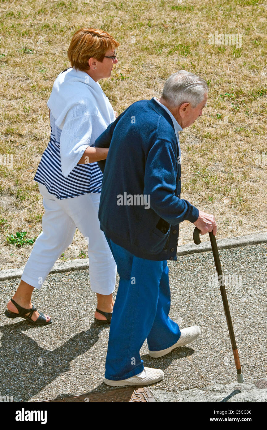Aider la femme homme plus âgé avec stick marche le long trottoir - France. Banque D'Images