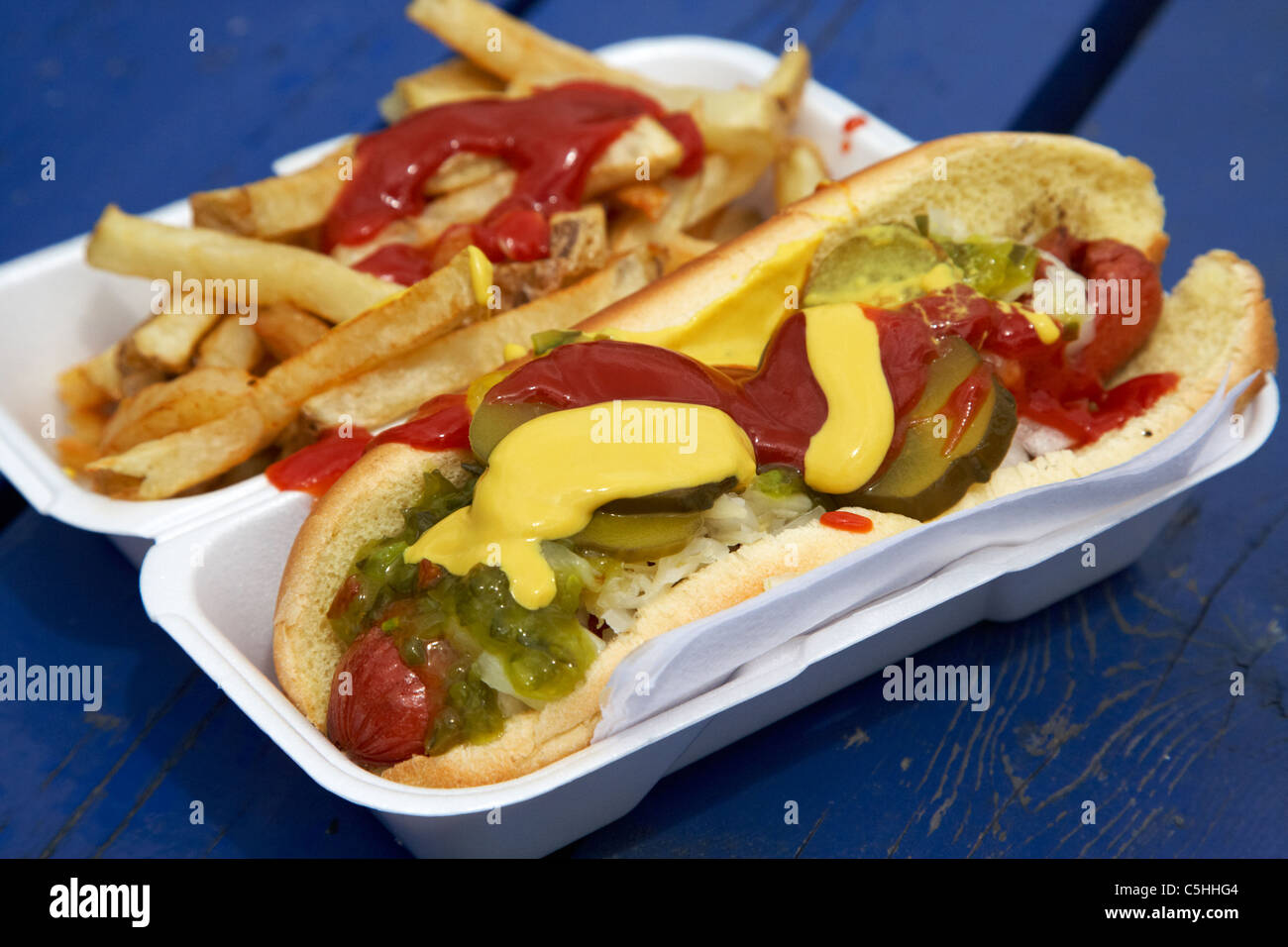 Hot-dog et frites à emporter couverts dans les sauces et condiments cornichons dans un récipient en polystyrène Banque D'Images