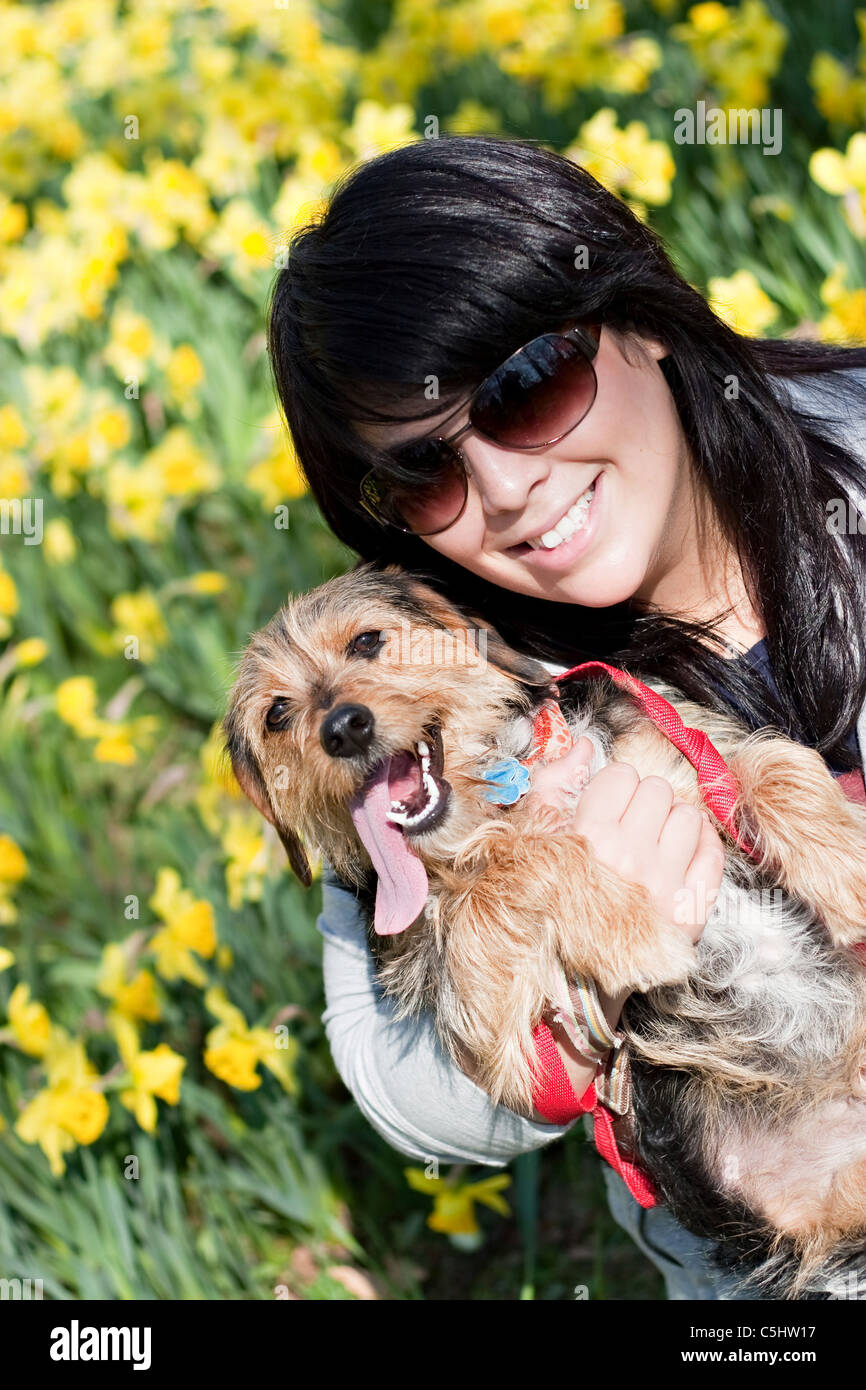 Une jeune femme tenant son chien de race mixte à l'extérieur au printemps en face d'un champ de fleurs de la jonquille. Banque D'Images