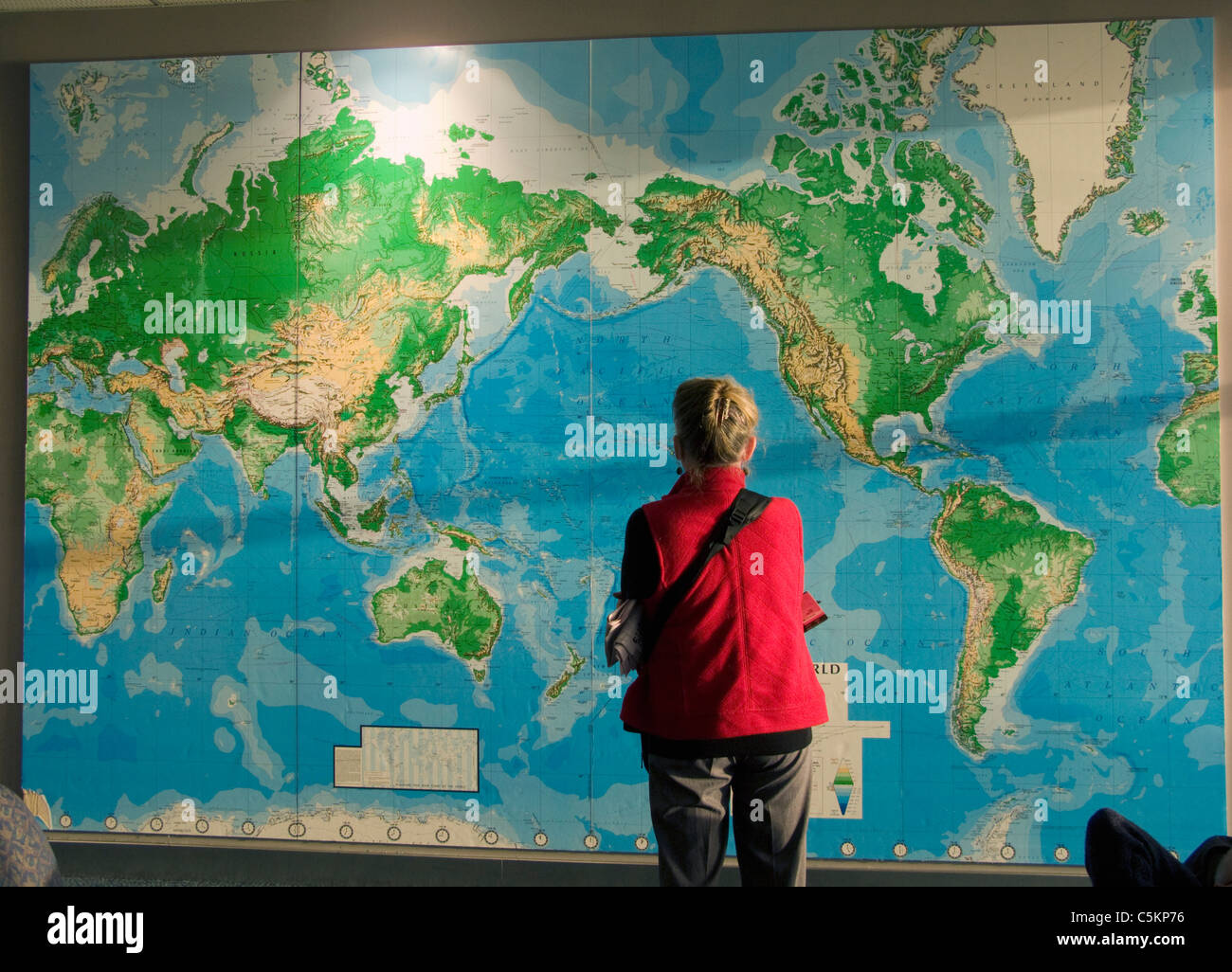 Une femme dans un manteau rouge, vue de dos, regardant une grande carte du monde sur un mur à l'aéroport d'Auckland Banque D'Images