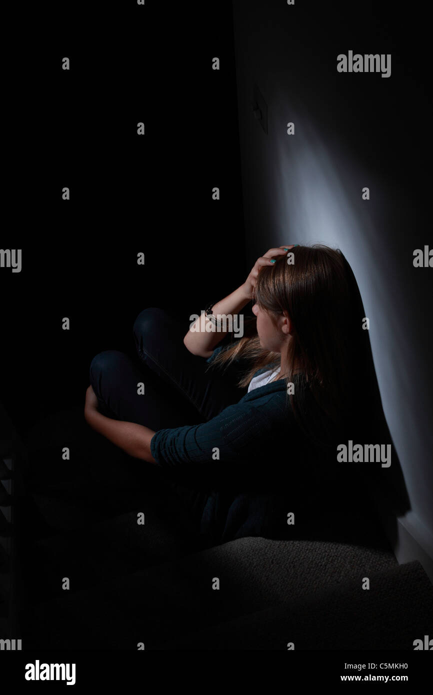 Jeune femme blonde assise seule dans le noir, la main sur la tête, vue de dos. Banque D'Images