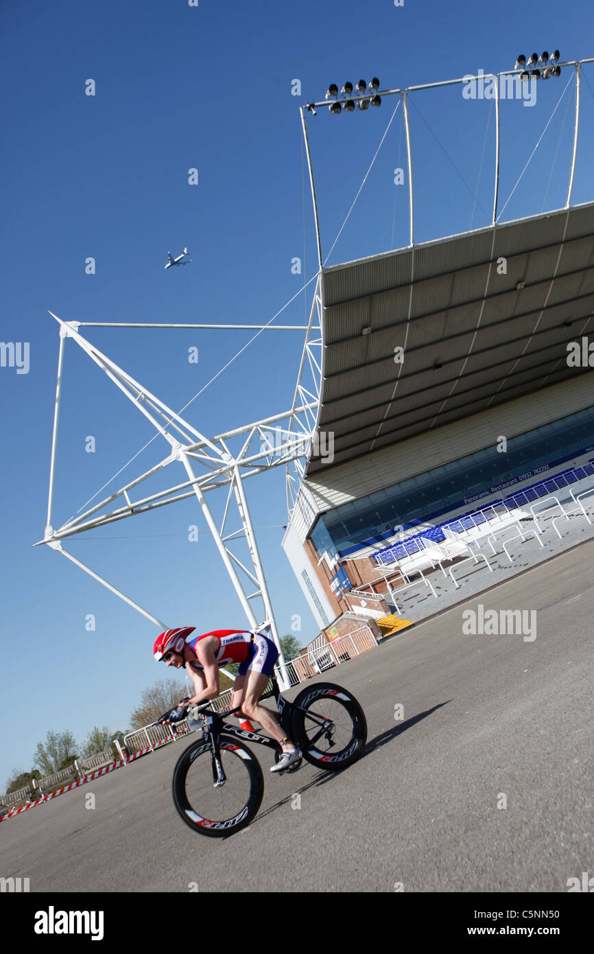 Un mâle concurrent cycliste passe Kempton Park Racecourse Road race lors d'une tribune dans un duathlon, et un avion vole par. Banque D'Images