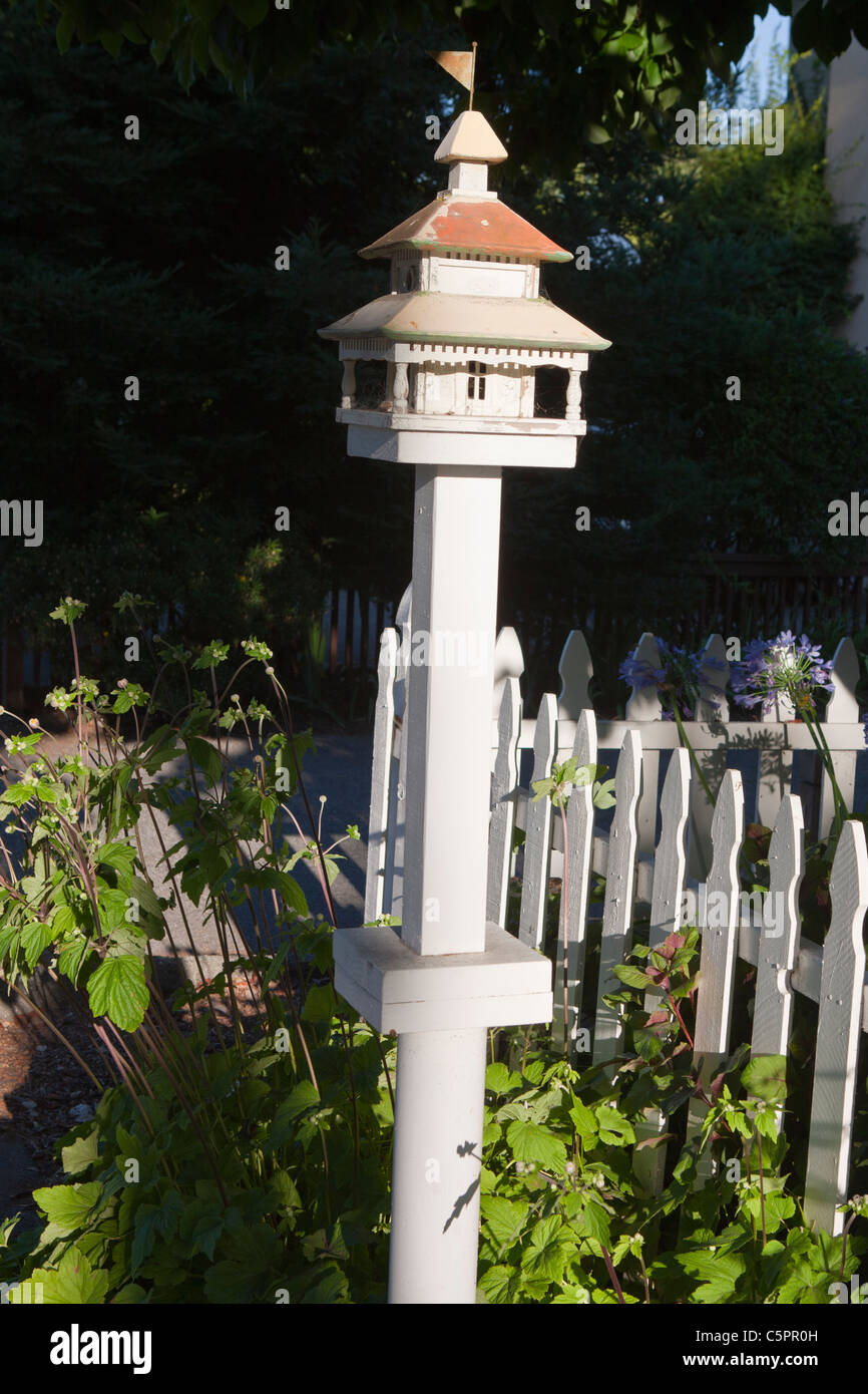 Maison d'oiseau blanc avec toit en métal et d'un drapeau en haut près de trottoir à côté de clôture blanche. Banque D'Images