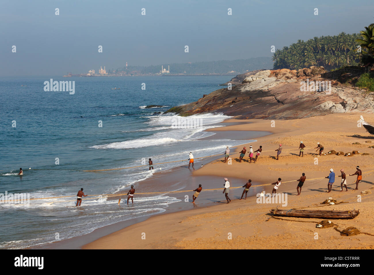 L'Inde, l'Inde du Sud, Kerala, côte de Malabar, pêcheurs tirant sur le net plage près de Kovalam, Vizhinjam en arrière-plan Banque D'Images