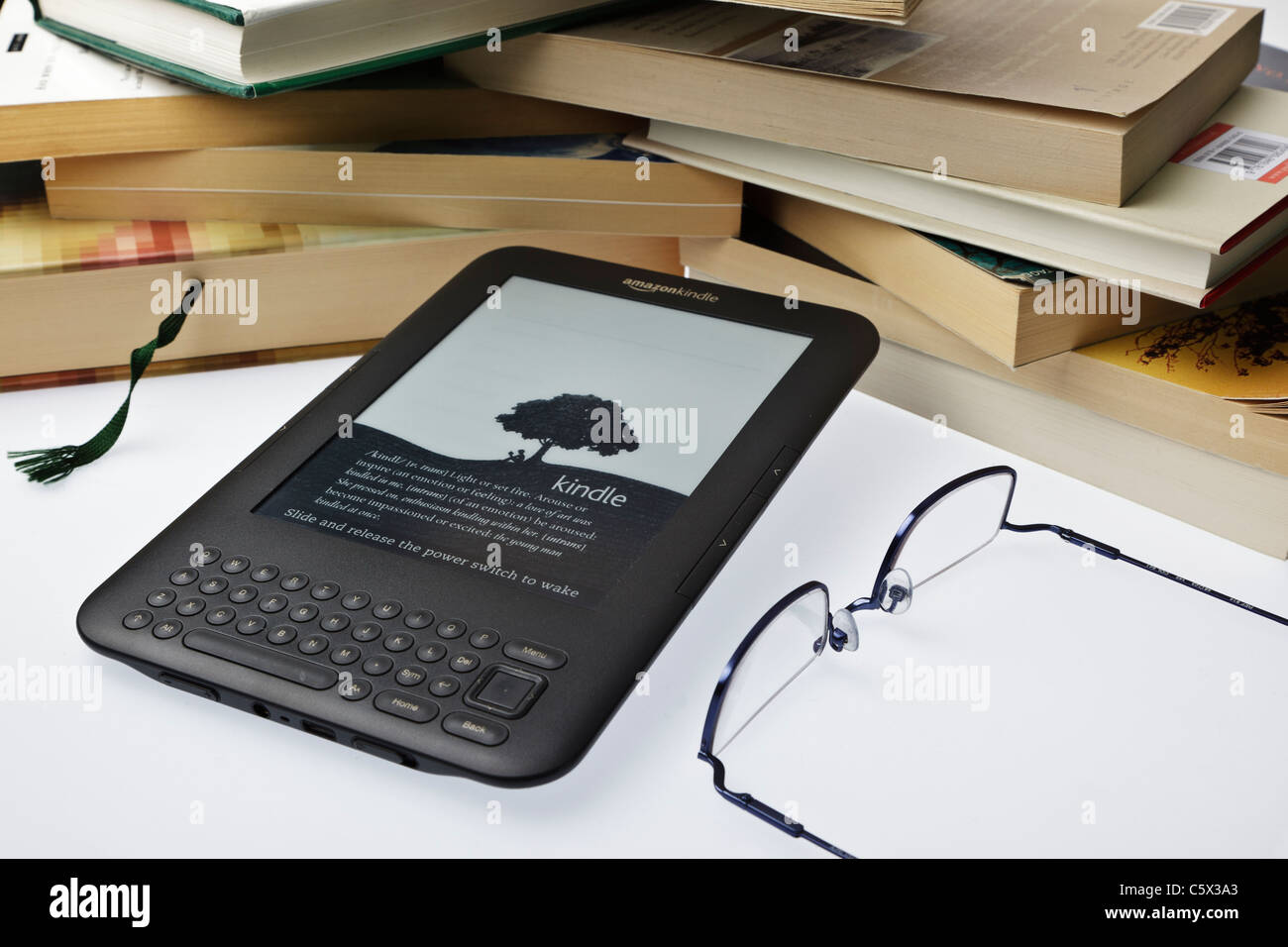 Amazon Kindle e-reader avec des lunettes de lecture et une pile de livres contre un arrière-plan uni, blanc Banque D'Images