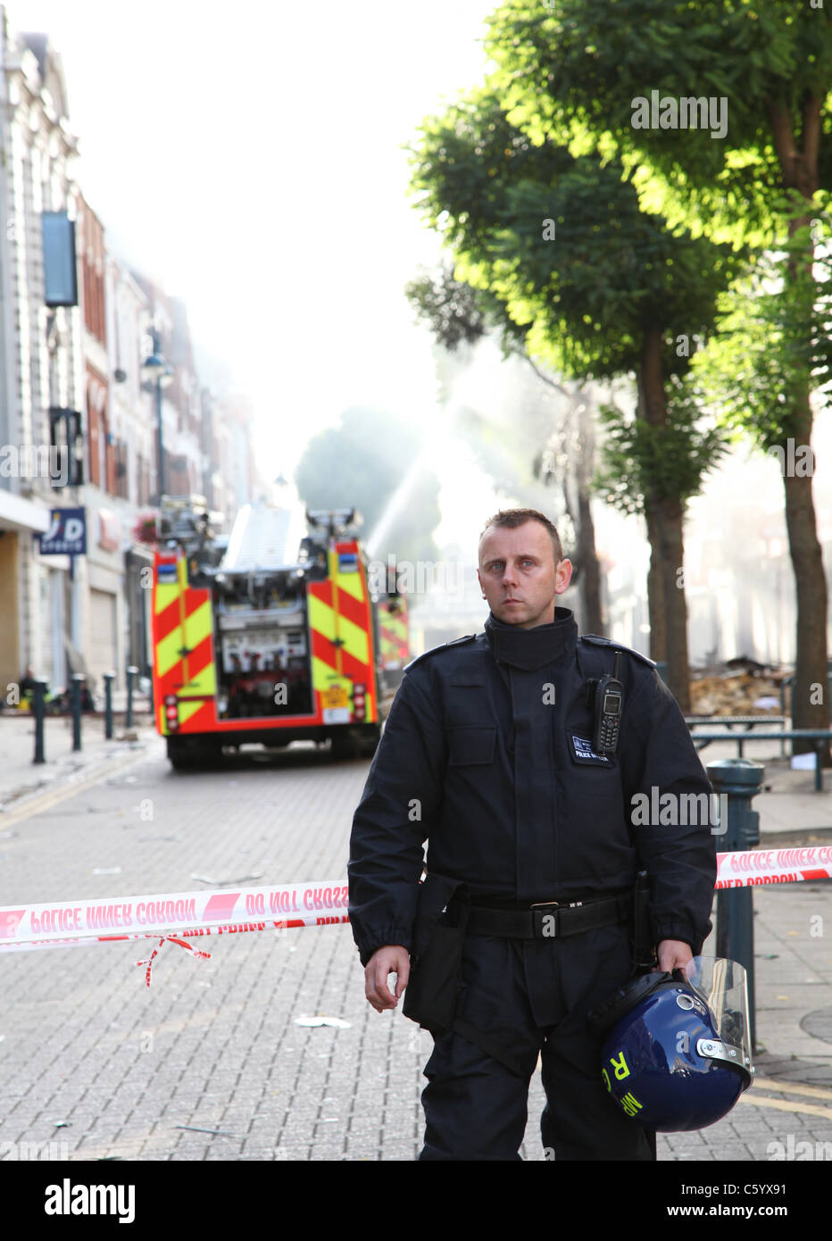 Agent de police anti-émeute avec le corned-off scène de crime, fire engine dans l'arrière-plan de la pulvérisation d'eau pour éteindre un feu Banque D'Images