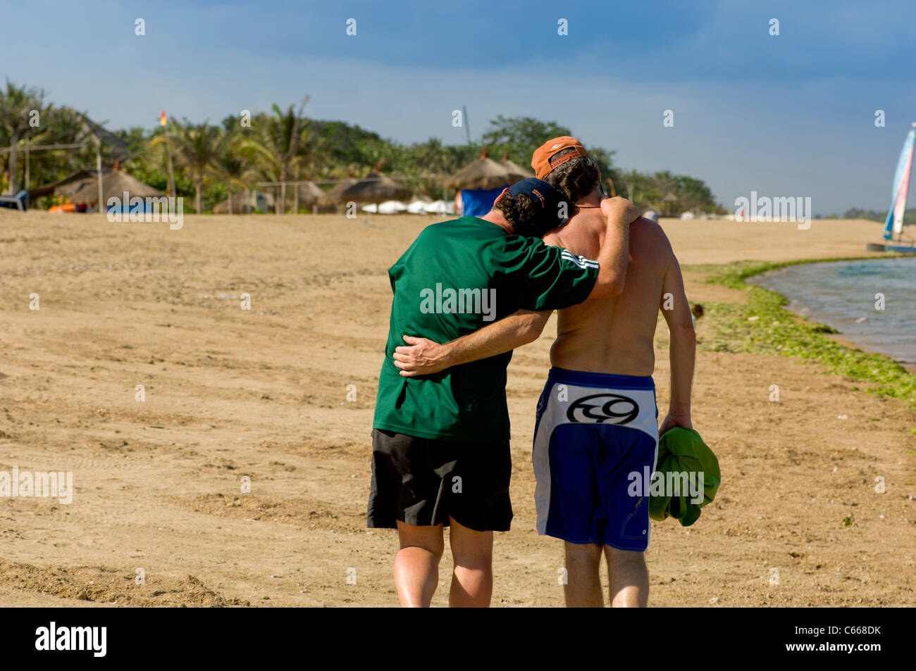 Vue arrière de deux hommes caucasiens portant un short marchant le long d'une plage avec des bras l'un autour de l'autre. Banque D'Images