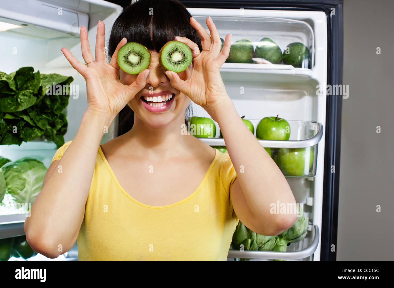 Mixed Race woman couvrant les yeux avec les tranches de kiwi près de réfrigérateur Banque D'Images Mixed Race woman couvrant les yeux avec les tranches de kiwi près de réfrigérateur Banque D'Images