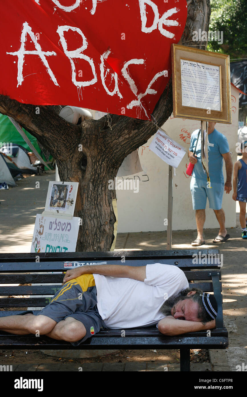 Une ville de tentes protestataire de dormir sur un banc de parc sous une bannière de 'stop' abus pendus dans le Boulevard Rothschild à Tel Aviv. Banque D'Images